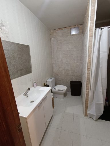 Baño pequeño con lavabo, inodoro y ducha con cortina blanca. Paredes de azulejos claros.