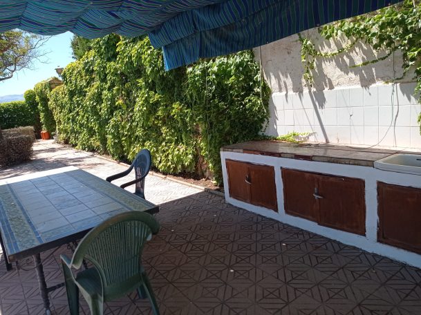 Área exterior con mesa, sillas y una zona de trabajo al aire libre rodeada de vegetación.