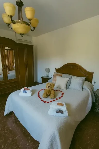 Habitación con cama, sábanas blancas, osito de peluche y toallas dispuestas en forma de corazón.