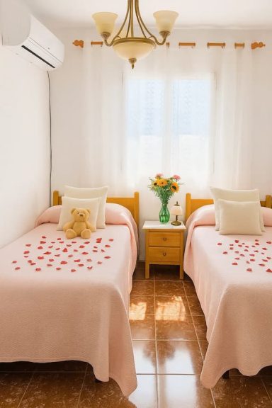 Habitación luminosa con dos camas, cojines y pétalos de rosa sobre las sábanas.