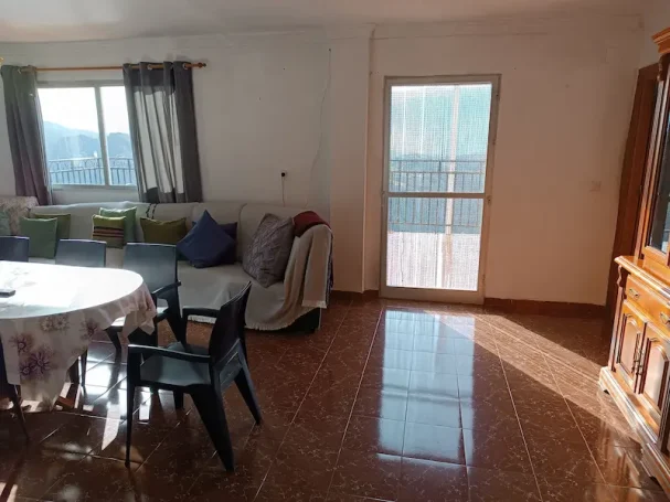 Sala iluminada con mesa, sillas, sofá y una puerta que da al exterior. Suelo brillante.