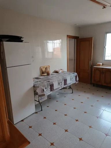 Cocina con nevera blanca, mesa con mantel y armario, paredes claras y suelo de baldosas.