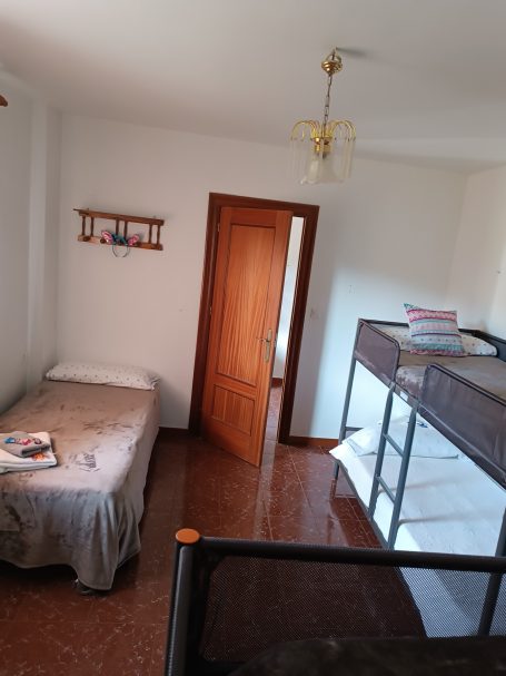 Habitación sencilla con cama individual, litera, estantería y puerta abierta.