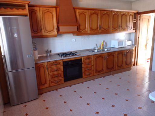 Cocina con muebles de madera, nevera, fogones y encimera clara. Suelo con baldosas.