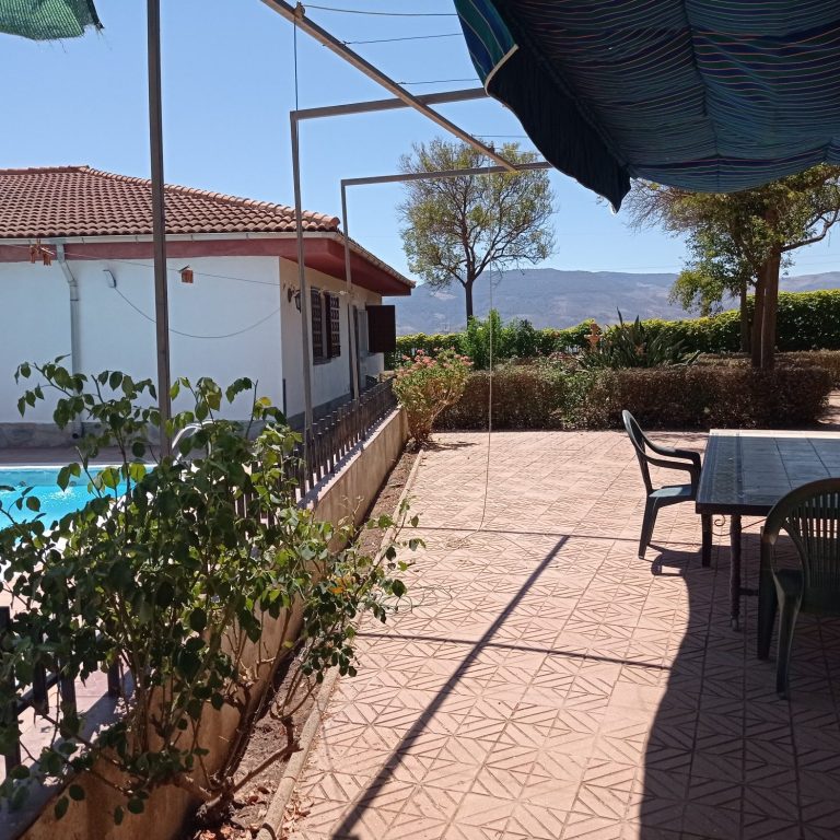 Terraza con mesa y sillas, piscina al fondo y montañas a lo lejos bajo un cielo despejado.