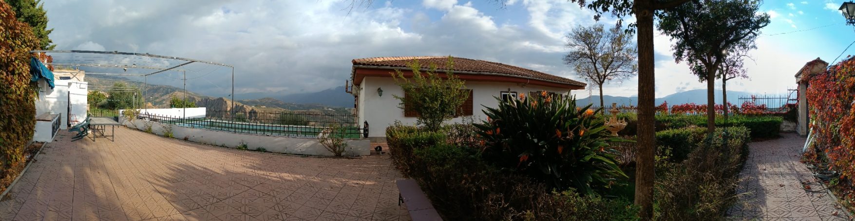 Vista panorámica de un jardín con piscina, casas y montañas al fondo.