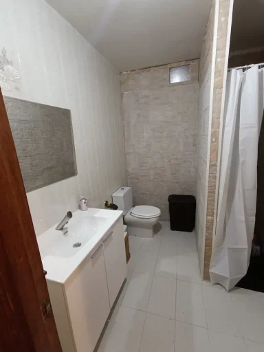 Baño con ducha, inodoro y paredes de azulejos claros. Ventana pequeña en la parte superior.