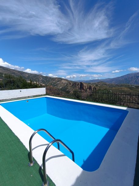 Piscina azul con vista a montañas y cielo con nubes en un día soleado.