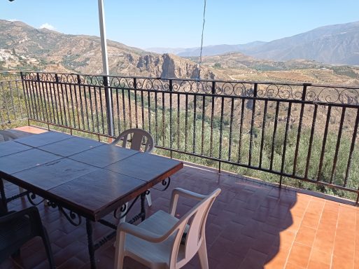 Mesa de madera con sillas en un balcón, vista de montañas y paisaje natural.
