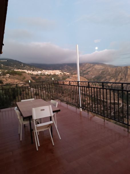Terraza con mesa y sillas, vistas a montañas y cielo nublado.