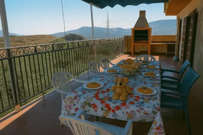 Mesa decorada con mantel floral, platos y comida, en un balcón con vistas montañosas.