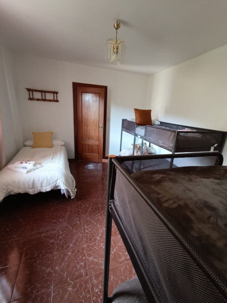 Habitación con una cama individual y una litera, paredes claras y suelo de cerámica.