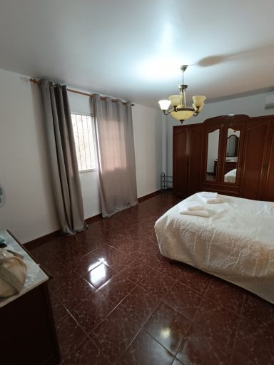 Habitación luminosa con cama, armario de madera y ventanas con cortinas. Suelo de cerámica.
