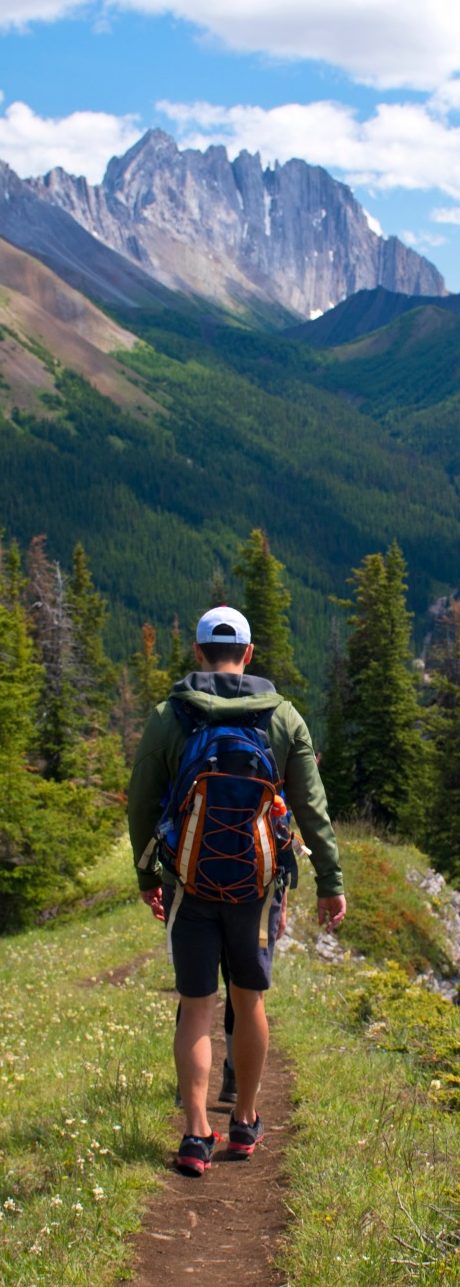 Hombre caminando por un sendero montañoso rodeado de árboles y montañas al fondo.