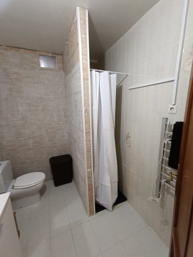 Baño con ducha, inodoro, lavabo y pared divisoria. Suelo y paredes de azulejos claros.