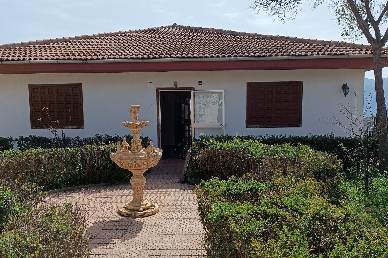 Fachada de una casa con una fuente en el jardín y ventanas con persianas.