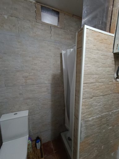 Baño con ducha, inodoro y paredes de azulejos claros. Ventana pequeña en la parte superior.