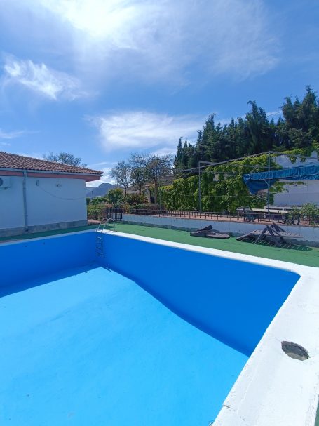 Piscina vacía con agua azul bajo un cielo soleado y vegetación alrededor.