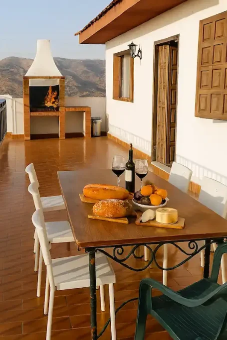 Mesa al aire libre con vino, pan y queso, vista de montaña y barbacoa en fondo.