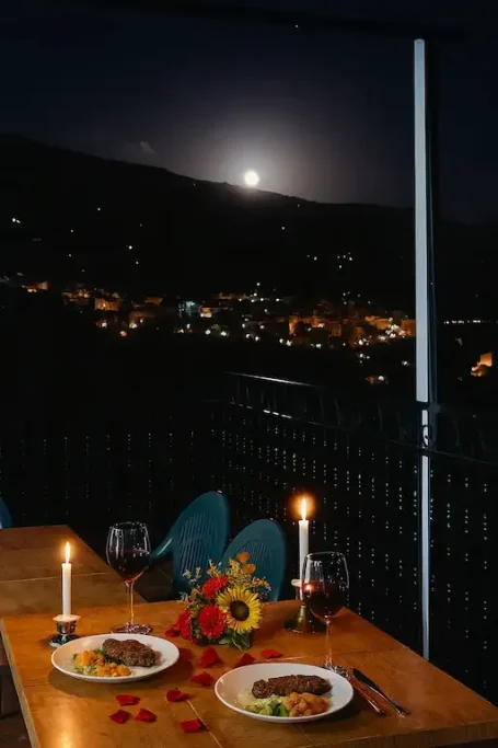 Mesa romántica con cena, velas encendidas y la luna llena sobre el paisaje nocturno.