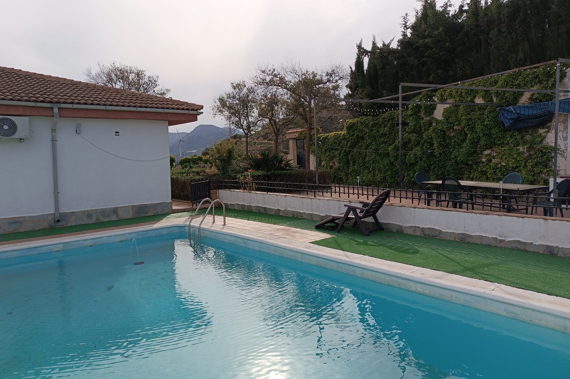 Piscina con borde de baldosas, rodeada de vegetación y un área de descanso.