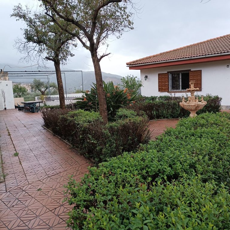 Jardín con árboles y plantas, alquiler casa blanca de fondo bajo un cielo nublado, Lanjarón