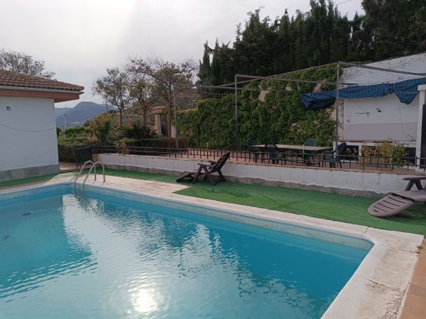 Piscina con jardín y montañas al fondo, rodeada de áreas de recreo y muebles exteriores.