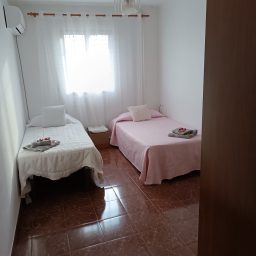 Habitación luminosa con dos camas, sábanas blancas y rosa, y ventanas cubiertas.